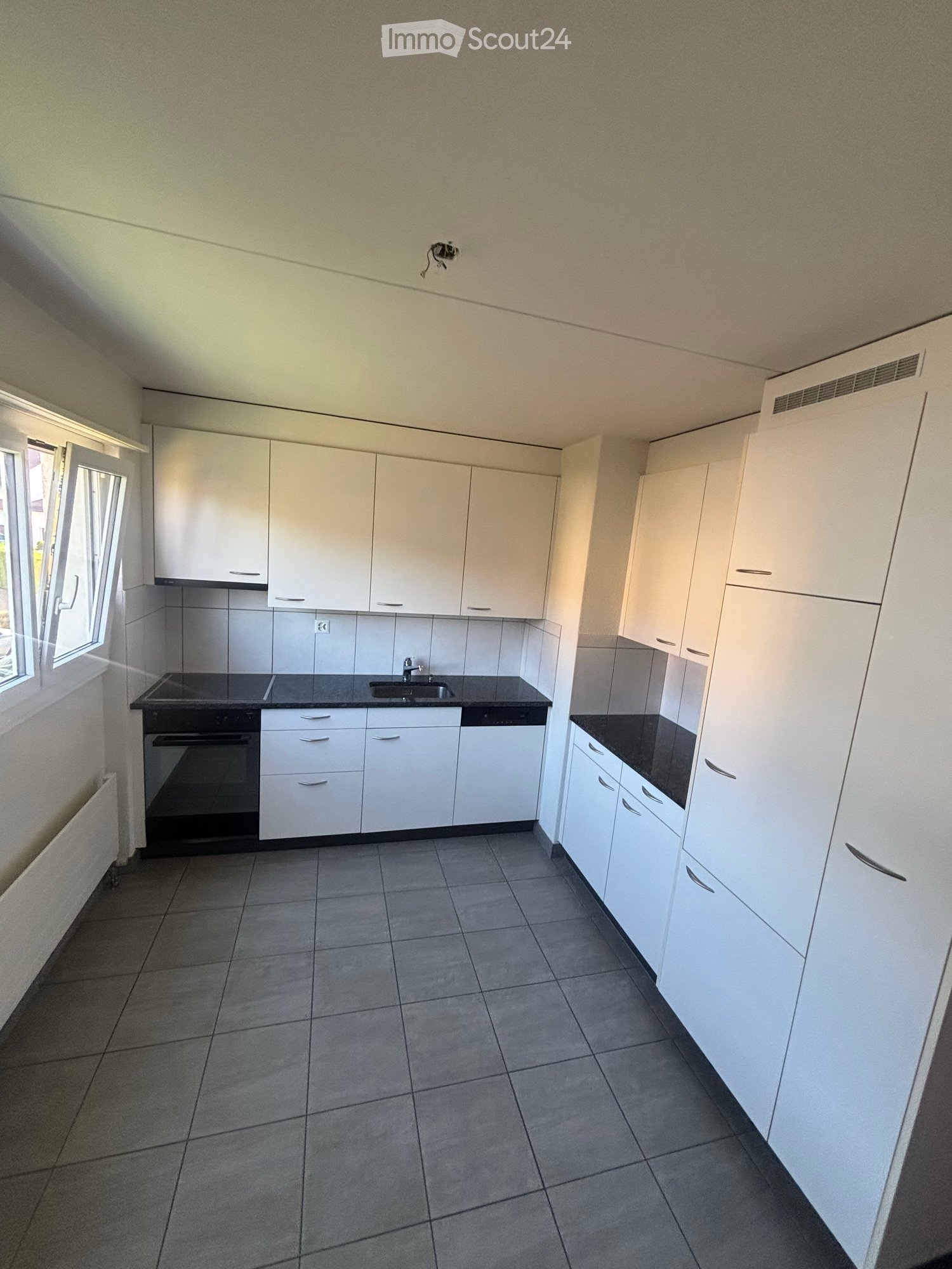 Empty kitchen with white cabinets, black countertops, and tiled flooring. Features include a sink, oven, and a window with blinds.