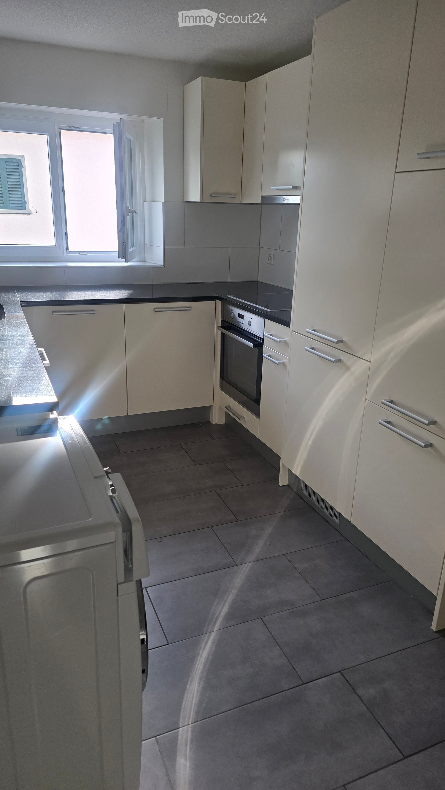 Modern kitchen with white cabinets, stainless steel oven, and dark tiled floor. Natural light streams in from a window on the left.