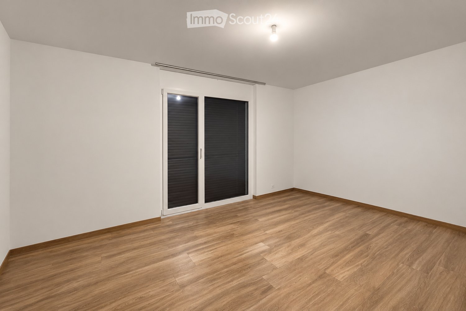 Empty room with wooden flooring, white walls, and large sliding glass doors with black blinds.