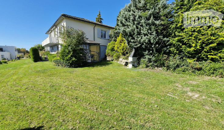 Well-maintained garden with a large, single-story house in the background. The house features a balcony, multiple windows, and a small shed. The garden is lush with various trees and shrubs, and a stone bench is visible near the house.