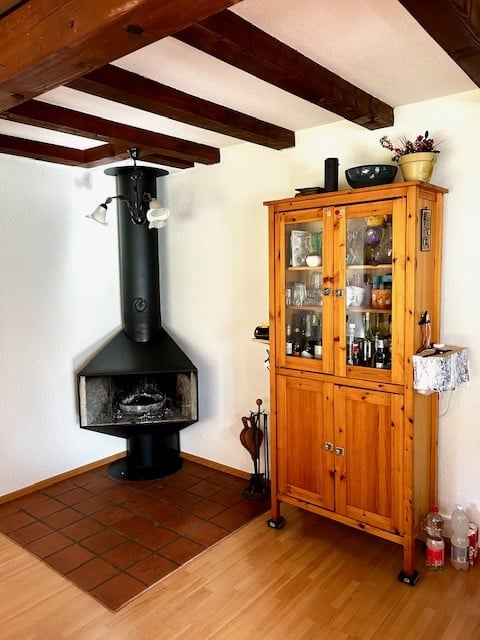 Cozy room with exposed wooden beams, a black wood-burning fireplace, a wooden display cabinet with glass doors, and a small vase on top.