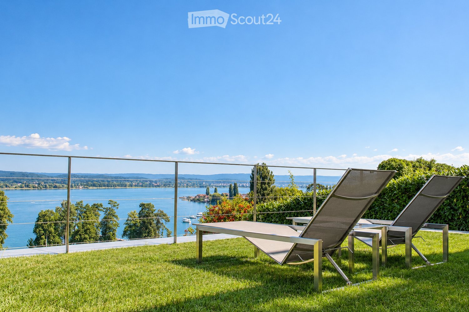 A modern outdoor patio with a metal table and two lounge chairs, overlooking a scenic lake and distant hills.