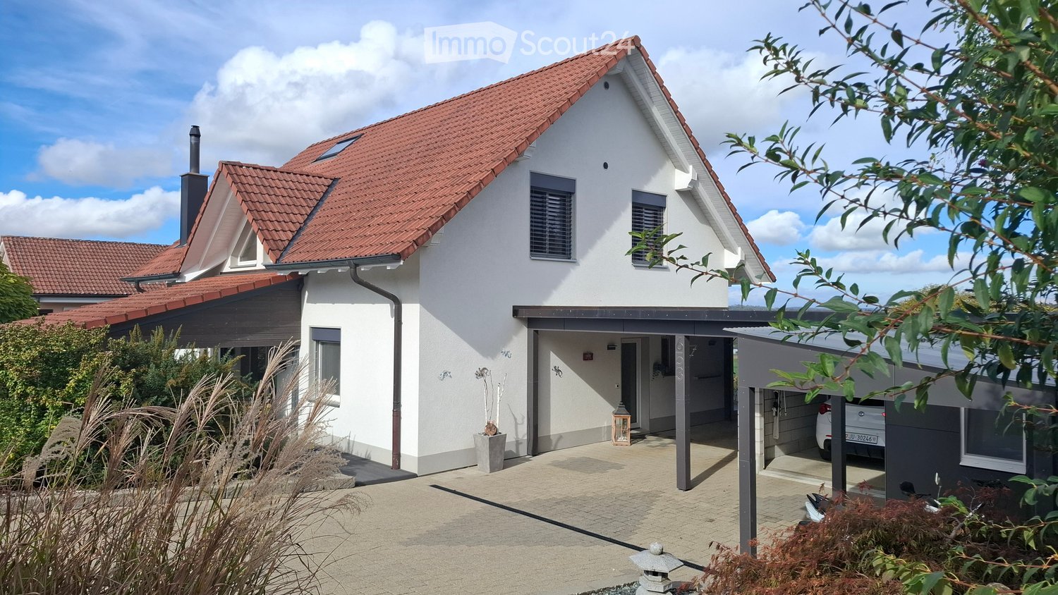 Modern two-story house with white exterior, red tile roof, and attached garage. Features include a covered entrance, small garden area, and a parked car in the garage.