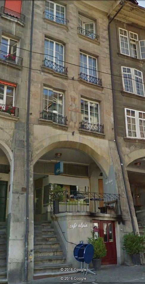 Multi-story building with arched entrance, balconies, and cafe signage. Classic European architecture with stone facade and decorative railings.