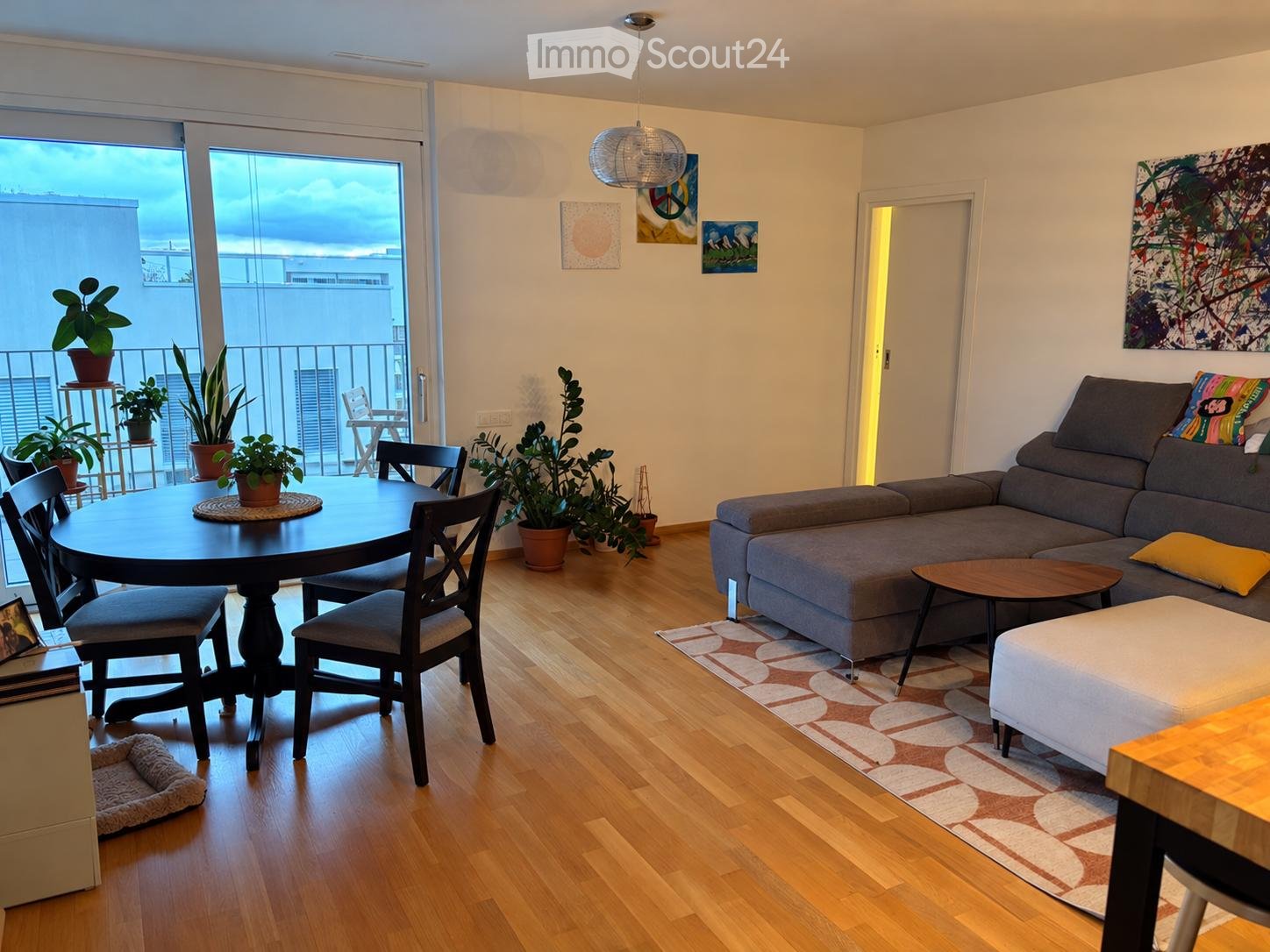 Bright living room with wooden flooring, large balcony access, grey sectional sofa, coffee table, ottoman, round dining table with chairs, potted plants, and a dog bed.