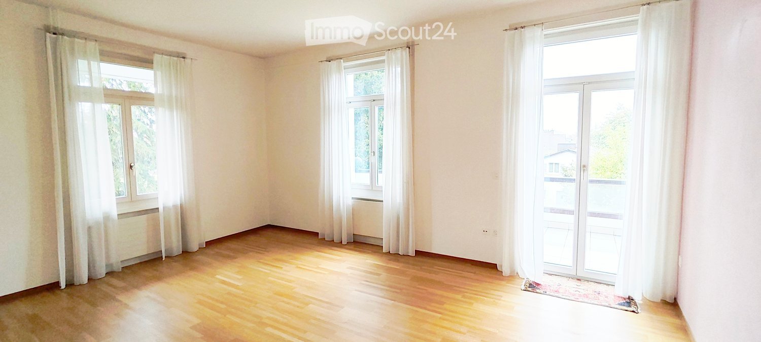 Empty room with wooden flooring, white walls, and large windows with white curtains. The room has a sliding glass door leading to a balcony.