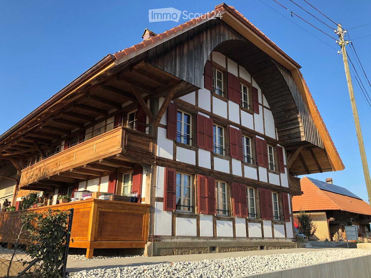 Traditional two-story wooden house with white and red exterior, multiple windows with shutters, and a wooden balcony on the upper floor.