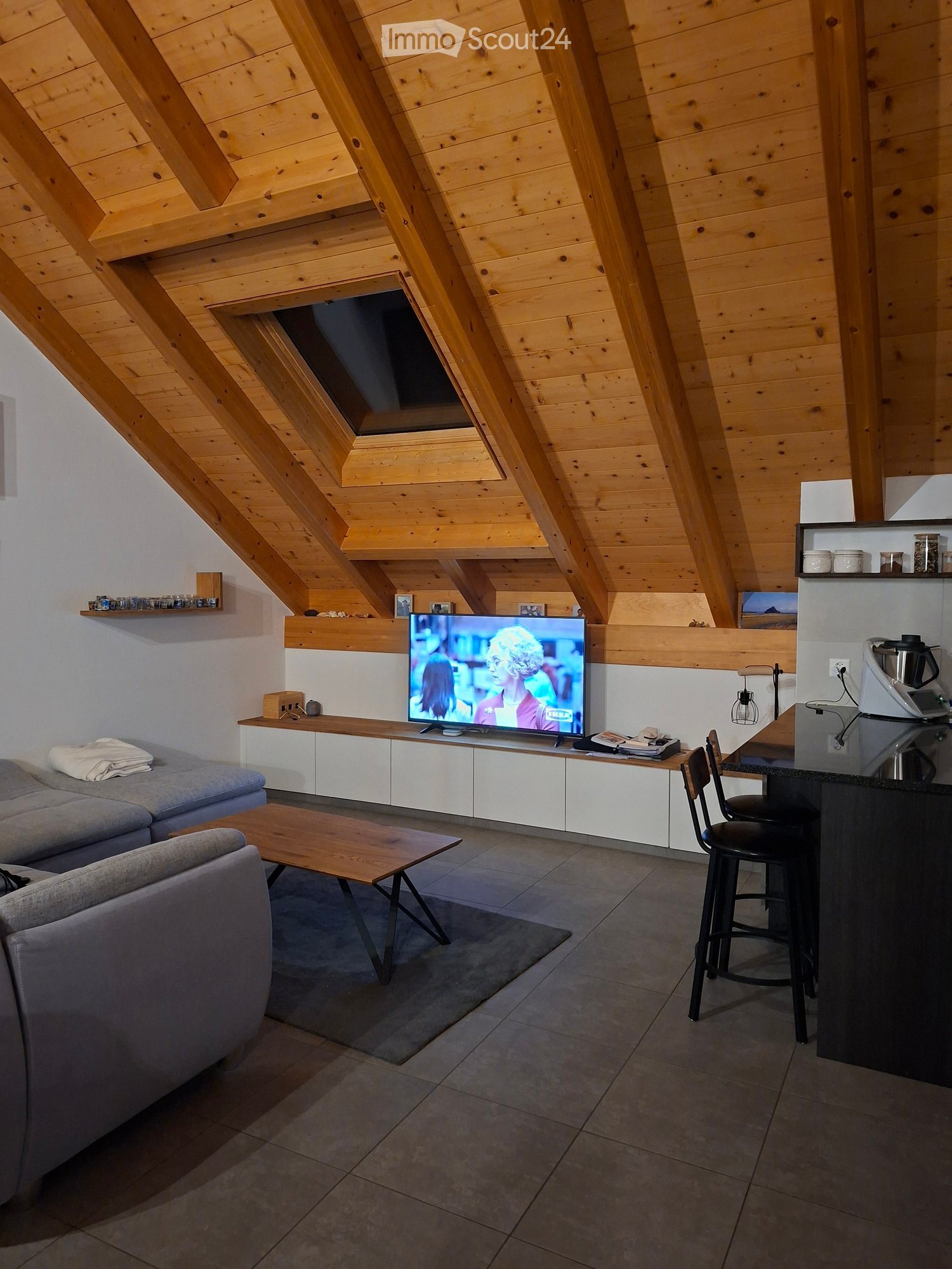 Modern living room with exposed wooden beams, grey sofa, coffee table, television, and kitchen counter with stools.