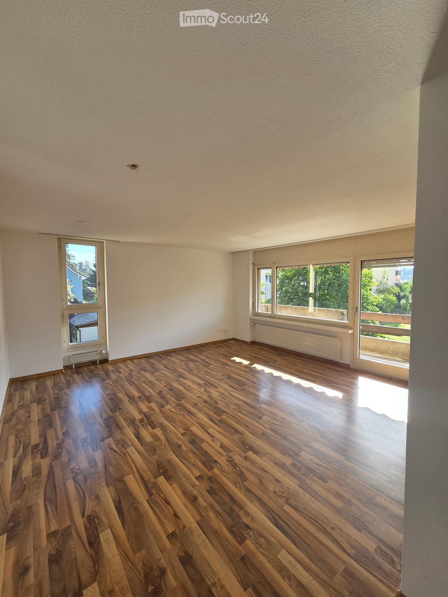 Empty room with wooden flooring, white walls, and large windows allowing natural light to enter.