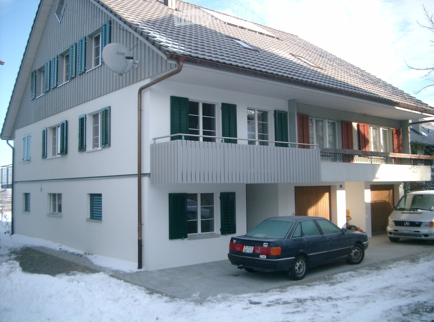 Two-story house with white exterior, green shutters, balcony on the upper floor, and a satellite dish on the roof. Two cars are parked in the driveway.