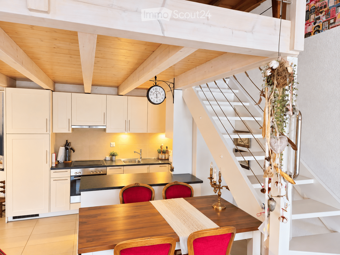Bright, modern kitchen with white cabinets, stainless steel appliances, and a wooden dining table with red chairs. A staircase leads to an upper level, decorated with a hanging floral arrangement and a candle holder on the steps.