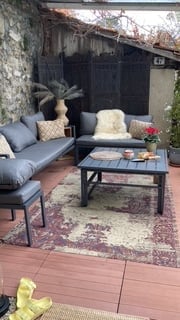 Outdoor patio with a grey sectional sofa, coffee table, patterned rug, and potted plants.