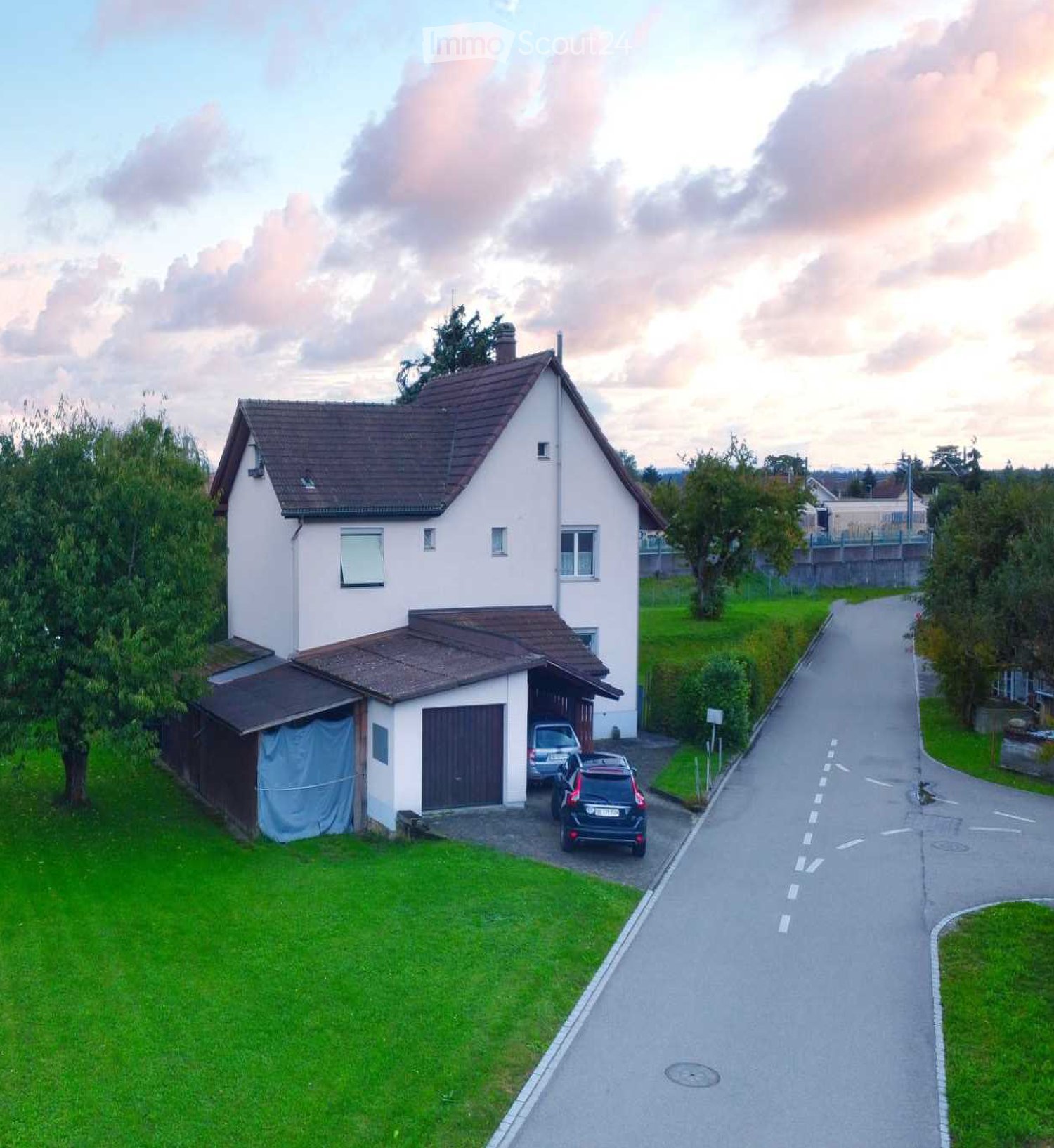 Two-story house with a brown tiled roof, white exterior walls, and a garage with a covered driveway. The house is surrounded by green grass and trees, with a paved road in front.