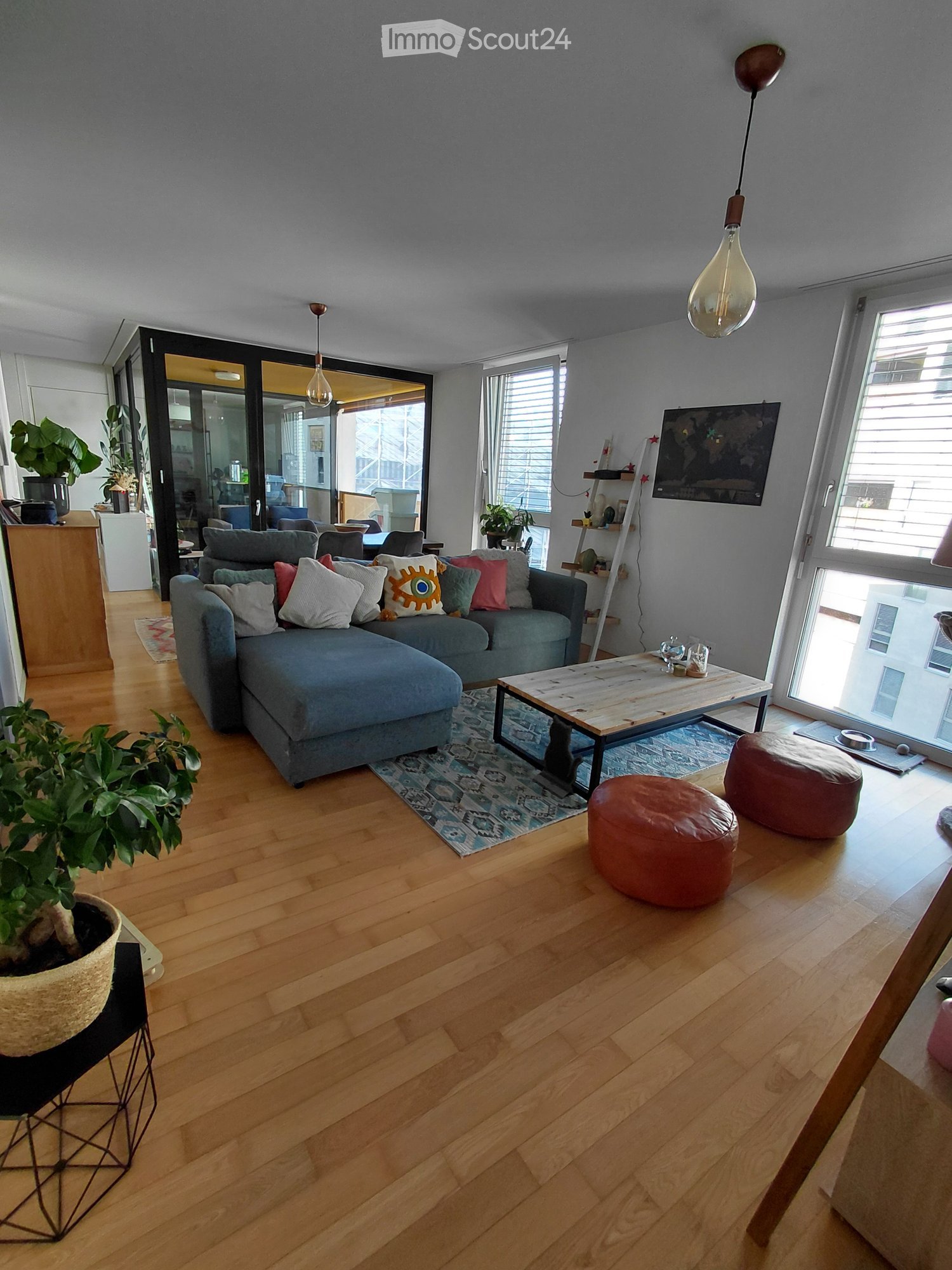 Modern living room with light wooden flooring, grey sectional sofa, colorful cushions, coffee table, ottomans, and potted plants.