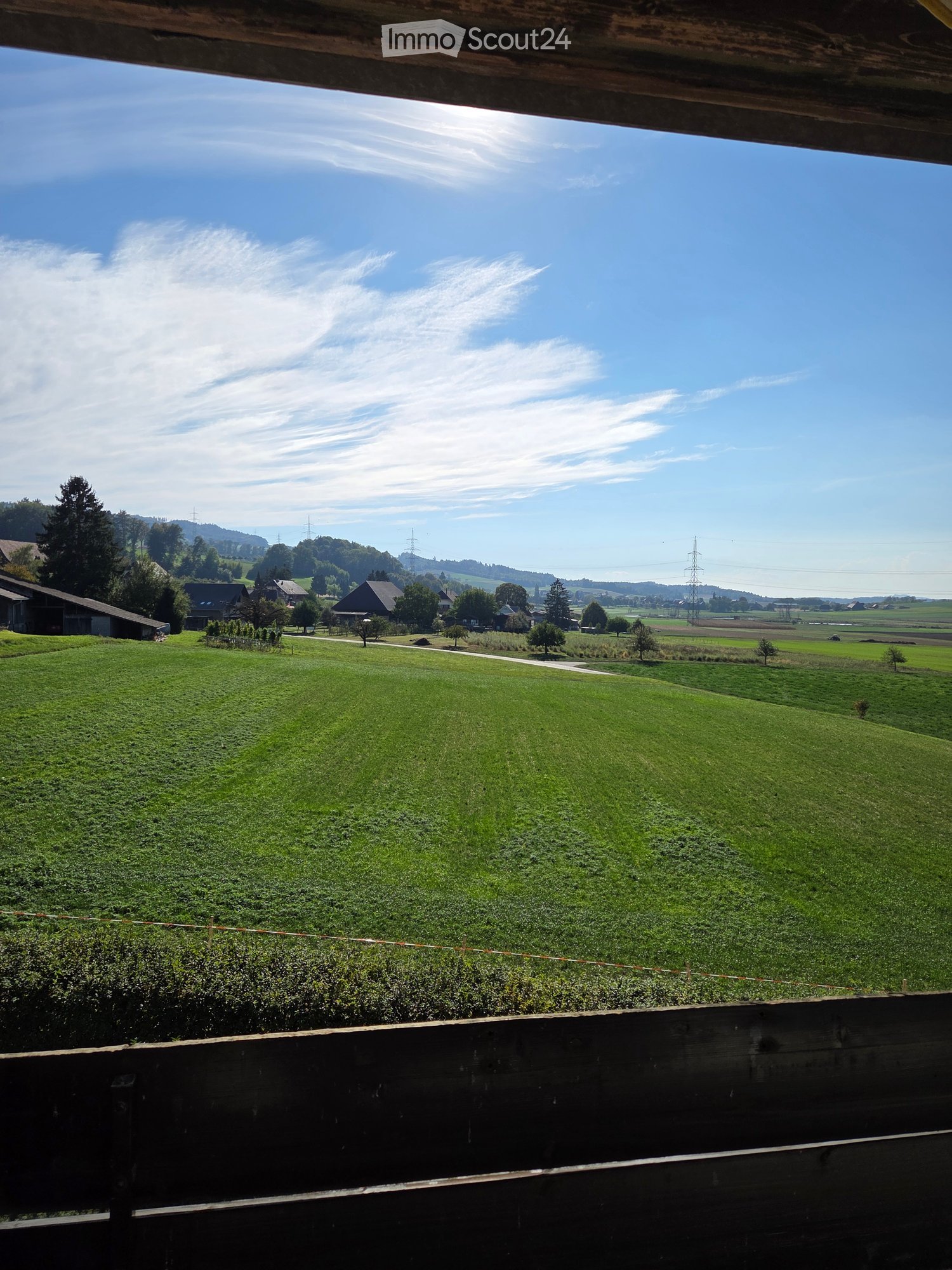 A panoramic view of a lush green field with scattered trees, distant houses, and a clear blue sky with scattered clouds.