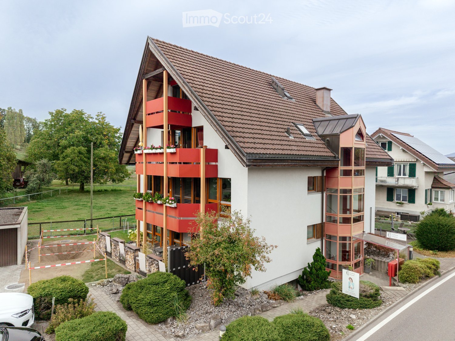 Modern two-story residential building with red and white exterior, multiple balconies, and surrounding greenery.