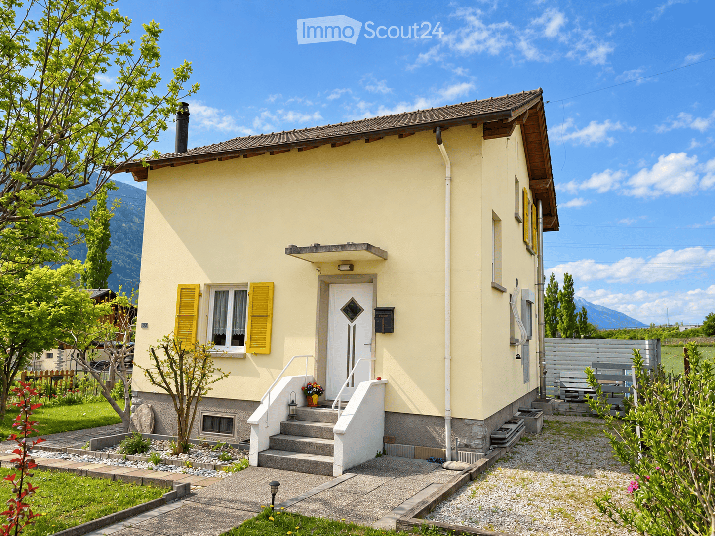 Single-story yellow house with white trim, raised ground floor, small porch, and garden area with a bench and plants.