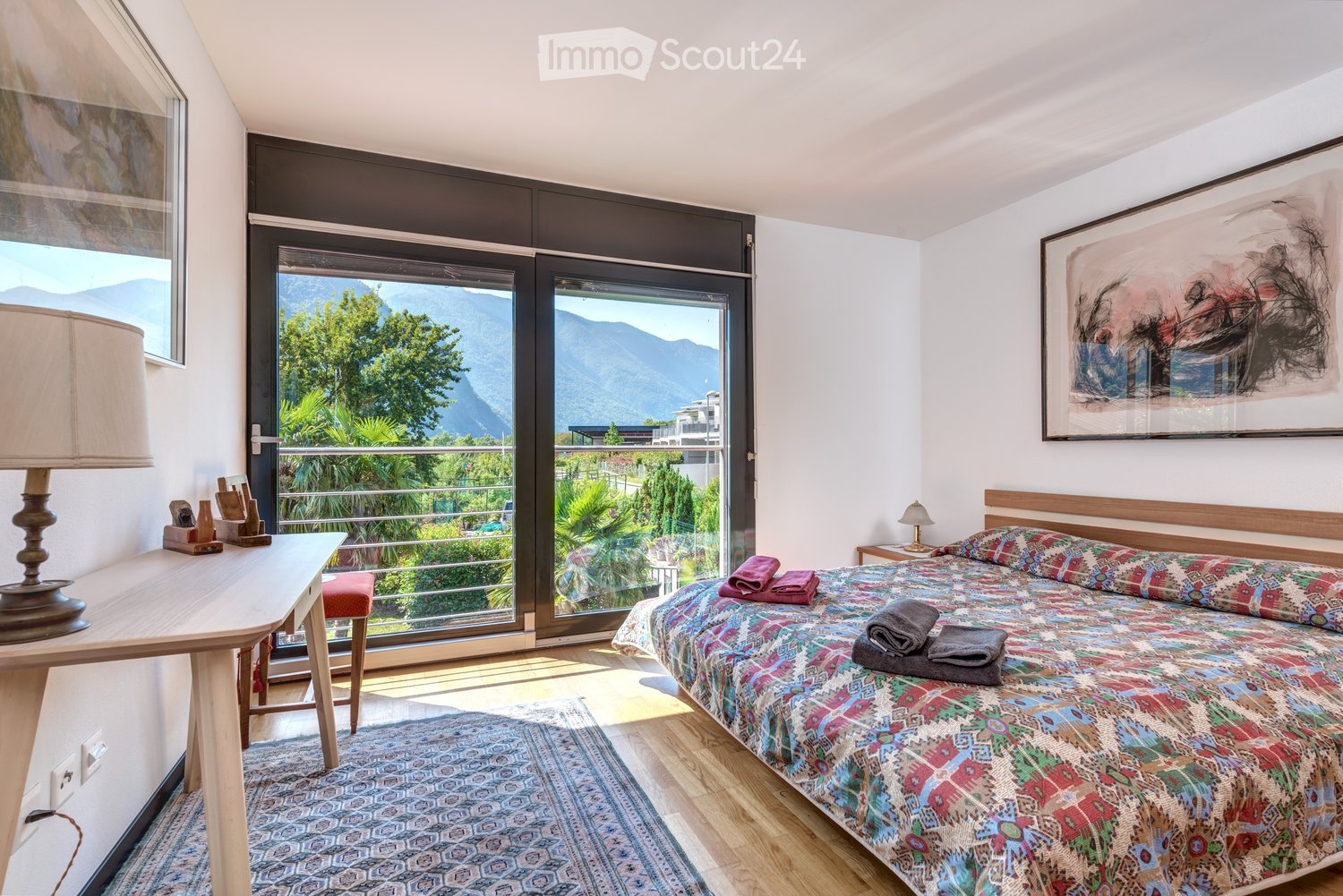 Bright bedroom with large windows offering a scenic mountain view, featuring a colorful bedspread, a wooden nightstand with a lamp, a small table with a lamp, and a framed artwork on the wall.