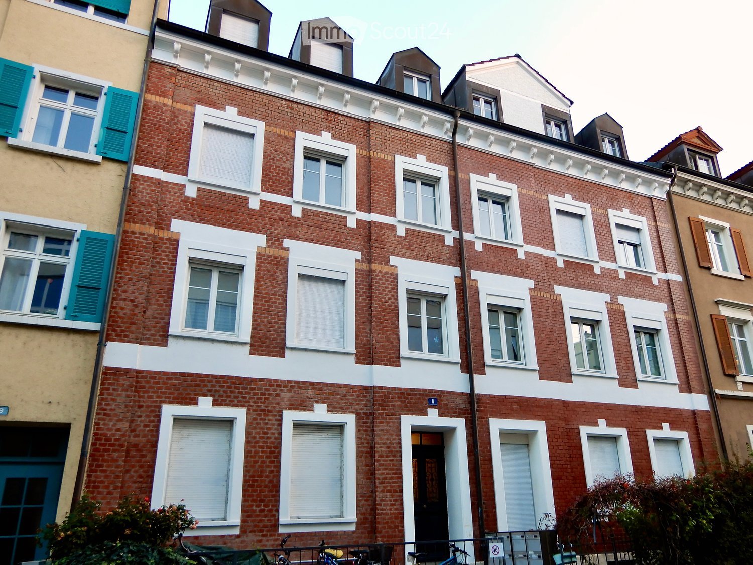 A multi-story brick building with white trim around windows and doors, featuring multiple windows with shutters and blinds, and a small balcony area with bicycles parked below.