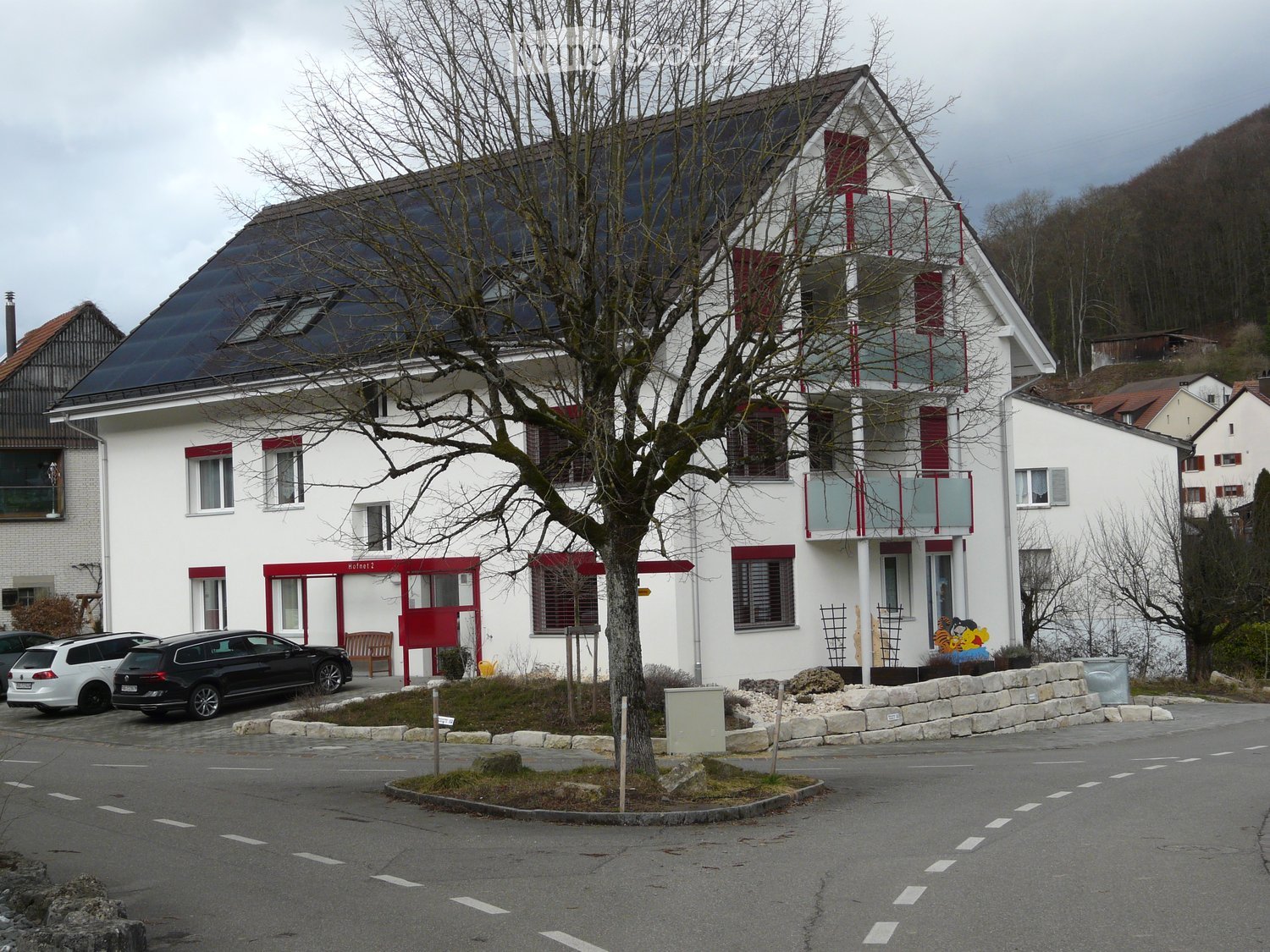 Two-story white building with red accents, large windows, and a balcony. Solar panels on the roof, parked cars, and a tree in front.