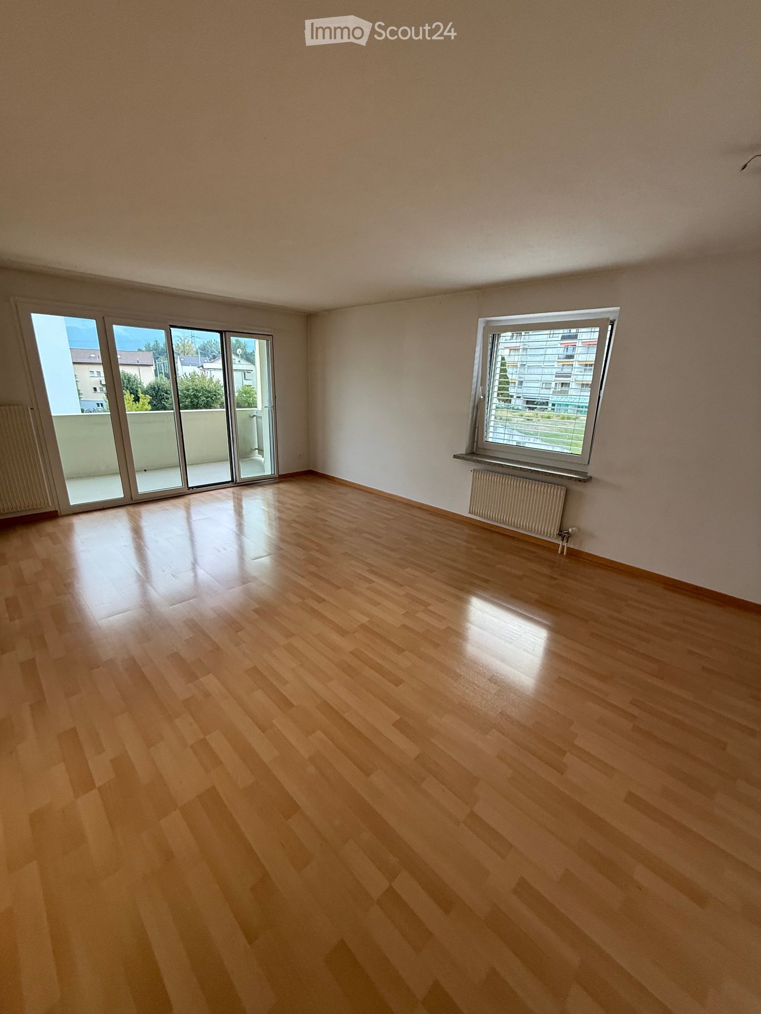 Empty room with wooden flooring, white walls, large windows, and sliding glass doors leading to a balcony.