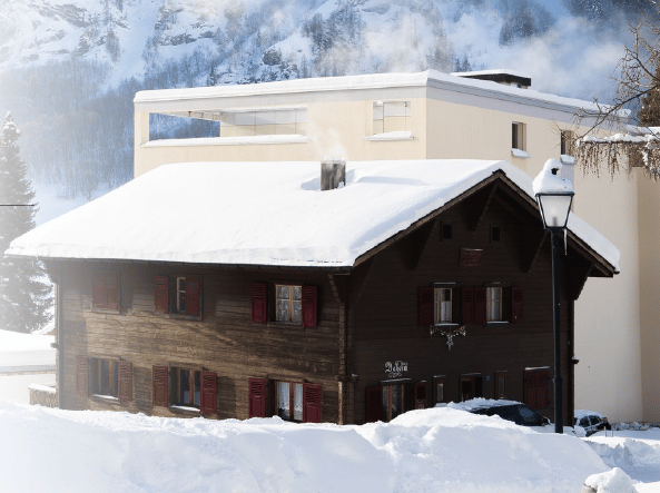 A traditional wooden chalet with red shutters and snow-covered roof, situated in a snowy mountain landscape with a modern building in the background.