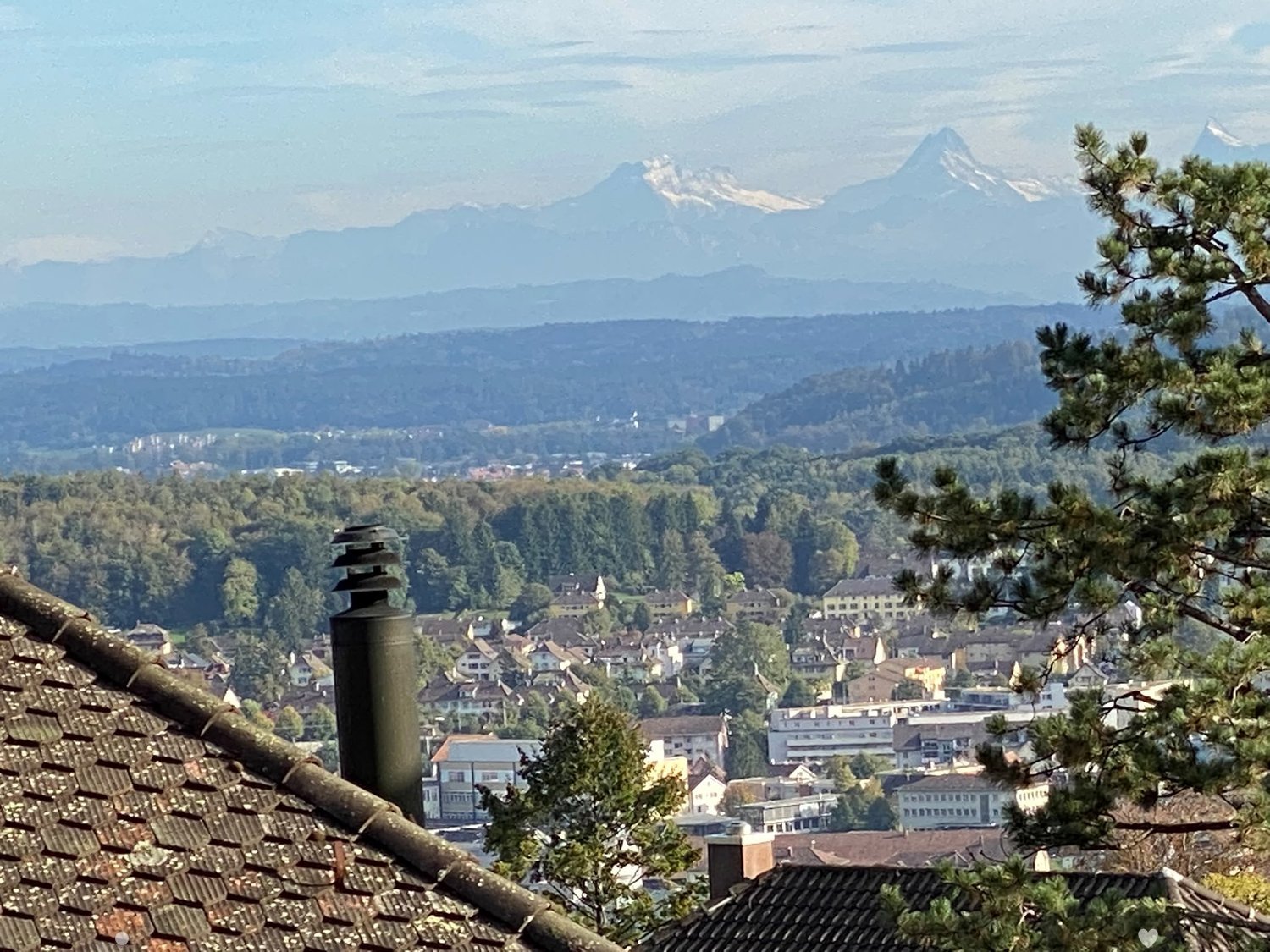 Panoramic view of a town with houses, trees, and mountains in the background.