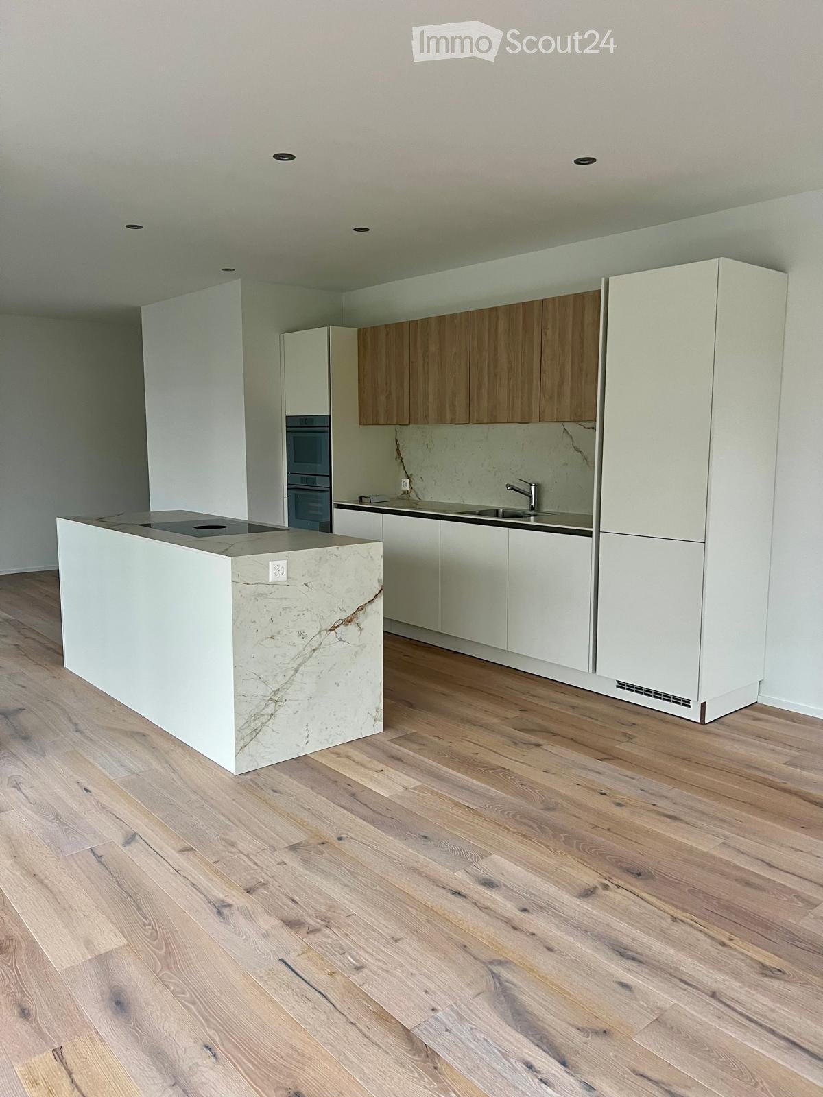 Modern kitchen with light wooden flooring, white and light wood cabinets, a central island with a marble countertop, and built-in appliances including an oven and sink.