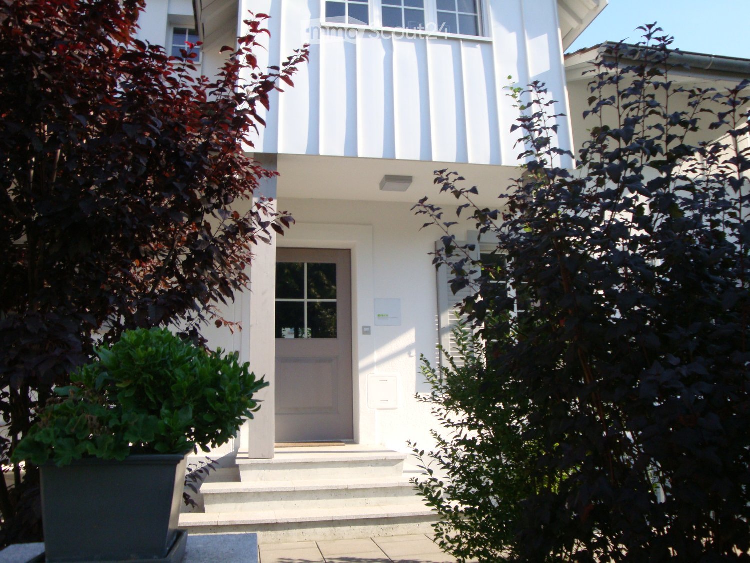 Two-story white house with a front door, steps, and surrounding greenery including bushes and trees.