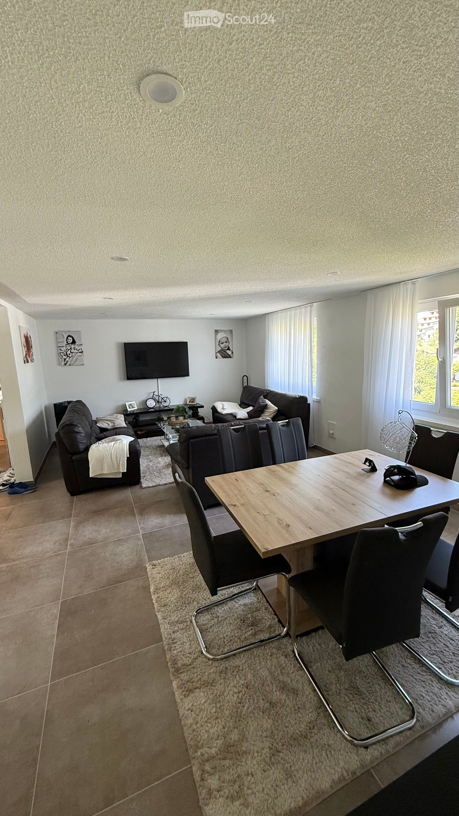 Modern living room with a sofa, coffee table, dining table, chairs, and a television mounted on the wall. The space features large windows with white curtains and a neutral color palette.