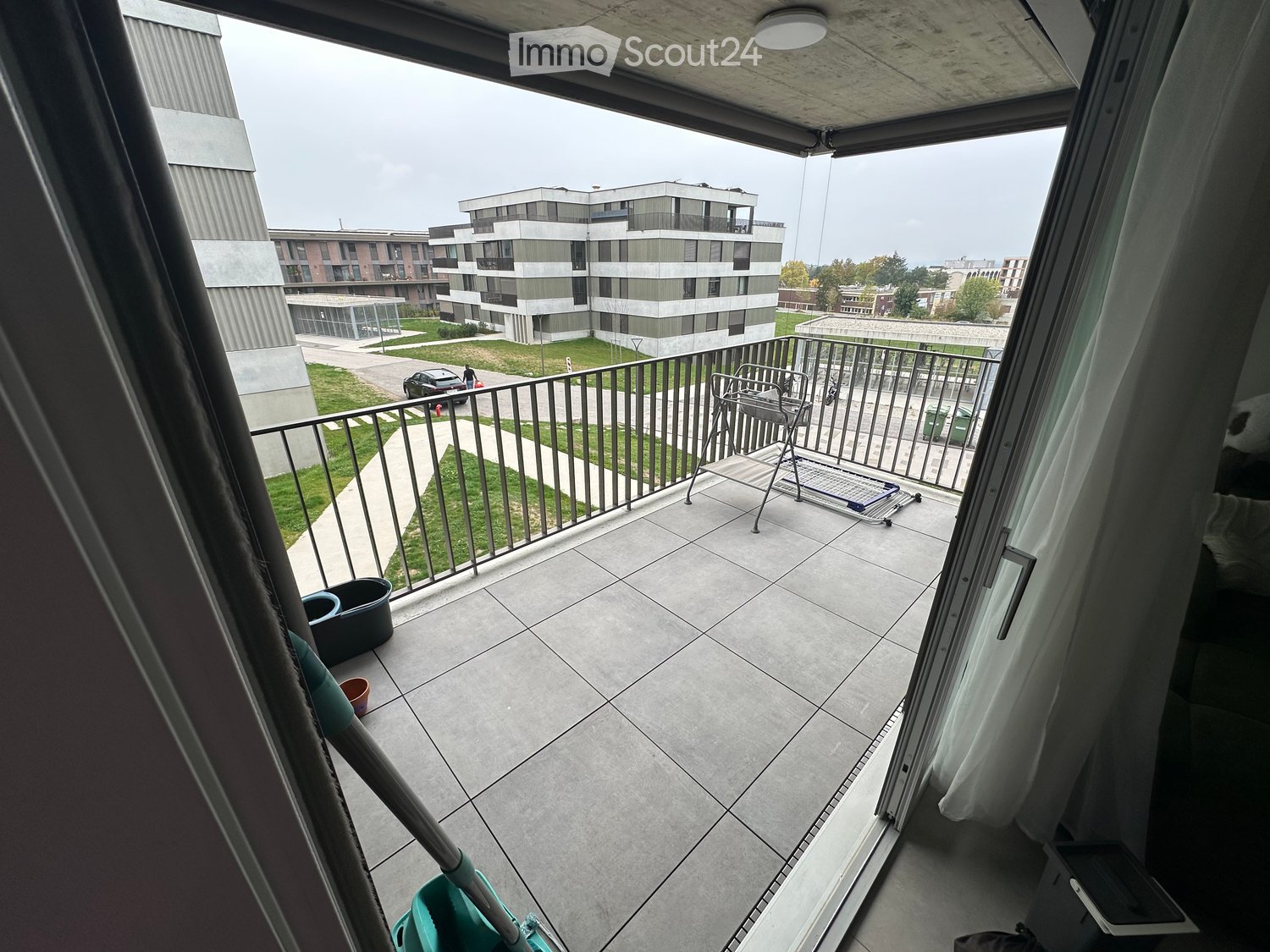 Balcony with tiled floor, metal railing, and a view of modern apartment buildings and a grassy area.