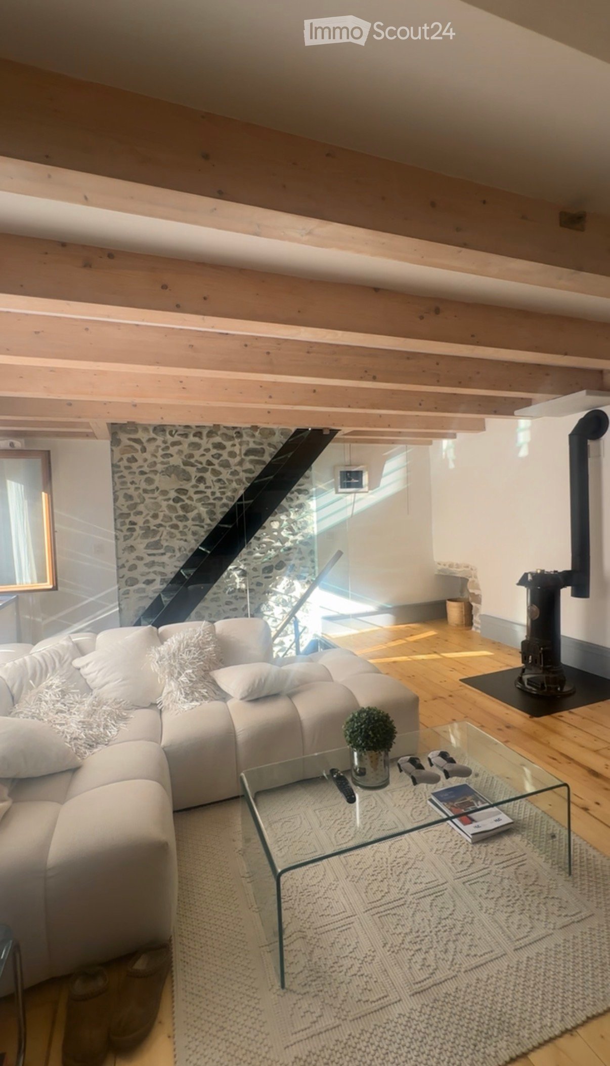 Modern living room with exposed wooden beams, a white sectional sofa, a glass coffee table, a small potted plant, and a fireplace. The room features a stone accent wall and a staircase leading to another level.
