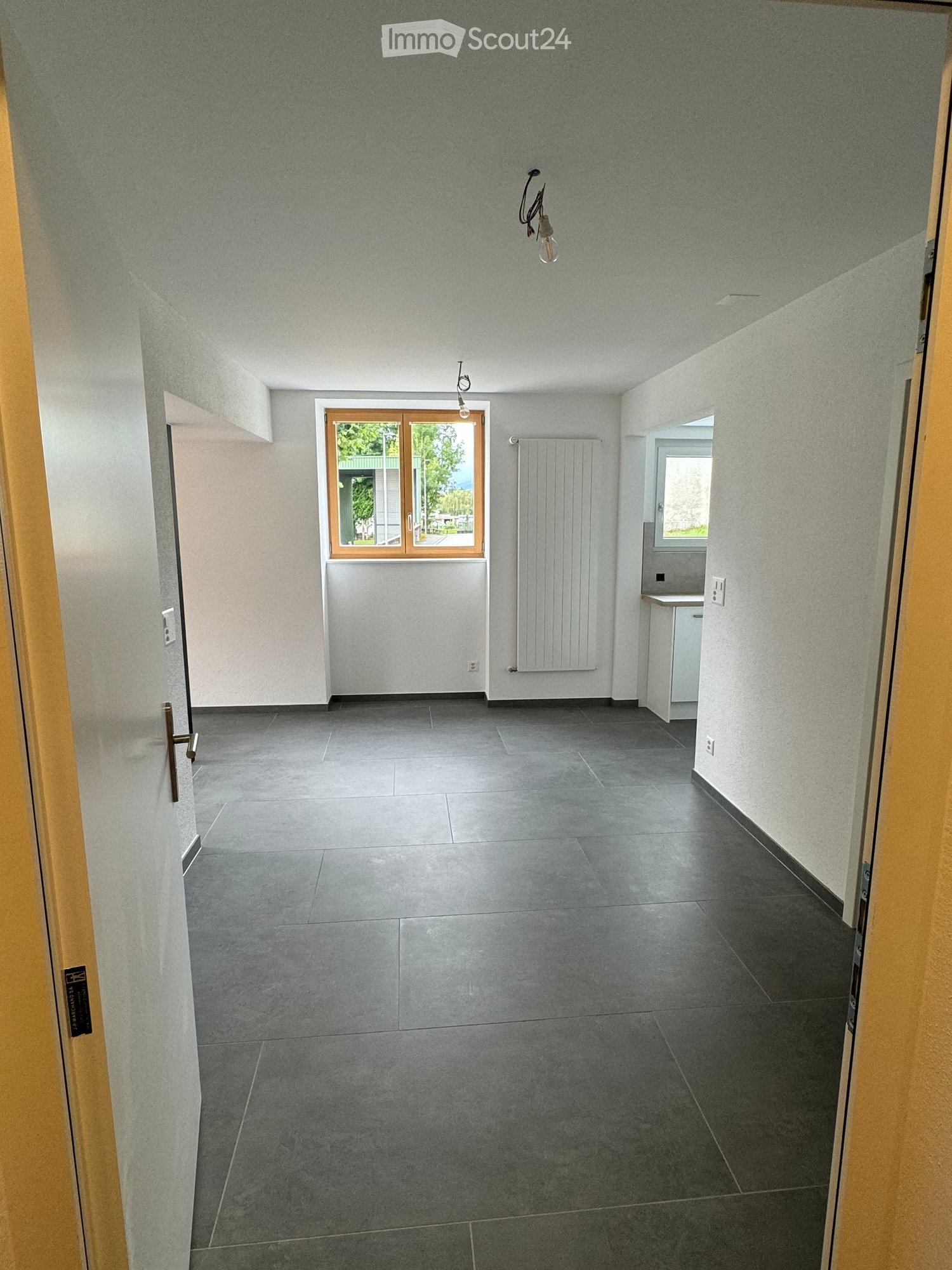 Empty room with grey tiled floor, white walls, and a window with wooden frame. There is a radiator and a small sink visible in the background.