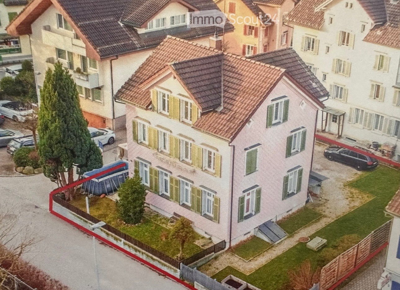 A two-story house with a red-tiled roof, multiple windows with shutters, and a small garden area with trees and a fence.