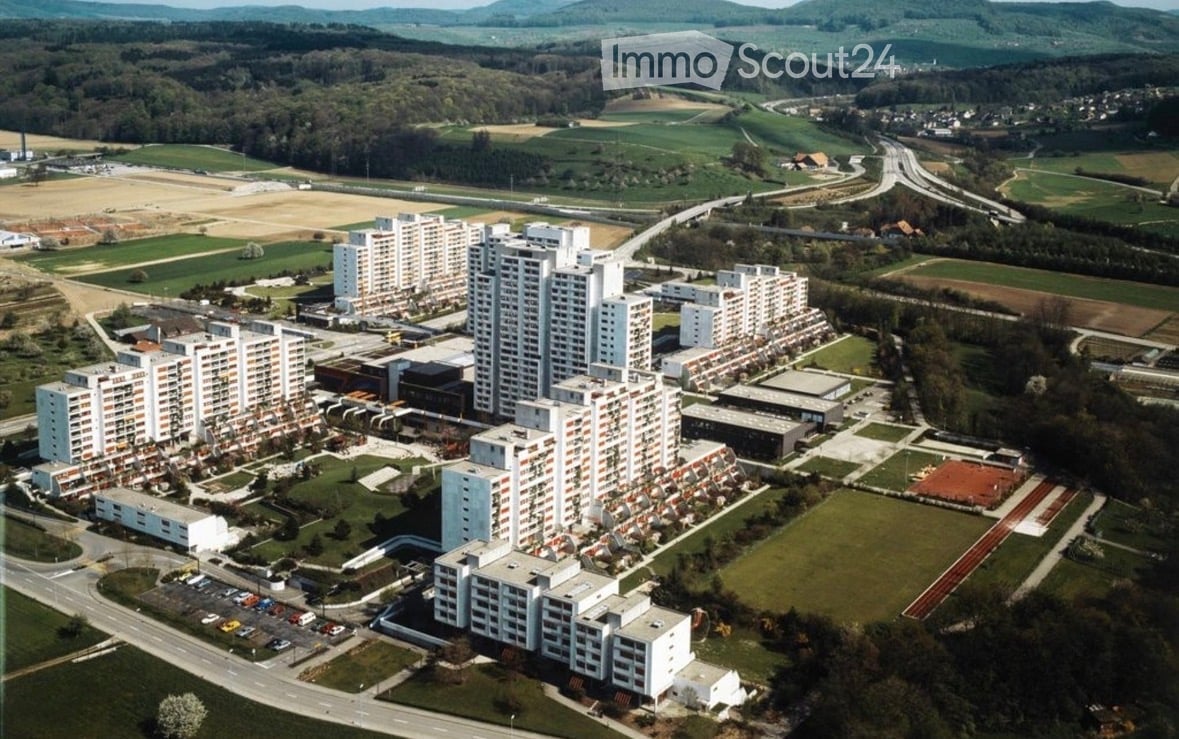 Aerial view of a large residential complex with multiple high-rise apartment buildings, surrounded by green spaces, roads, and distant hills.
