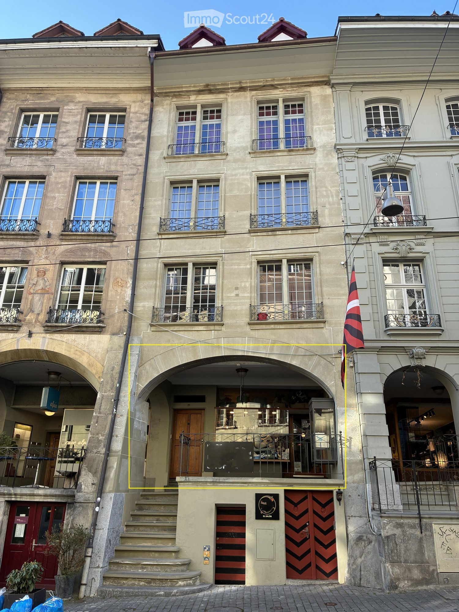 A multi-story building with ornate architecture, featuring arched entrances, balconies with metal railings, and large windows. The facade includes decorative stonework and a flag displayed on the right side.