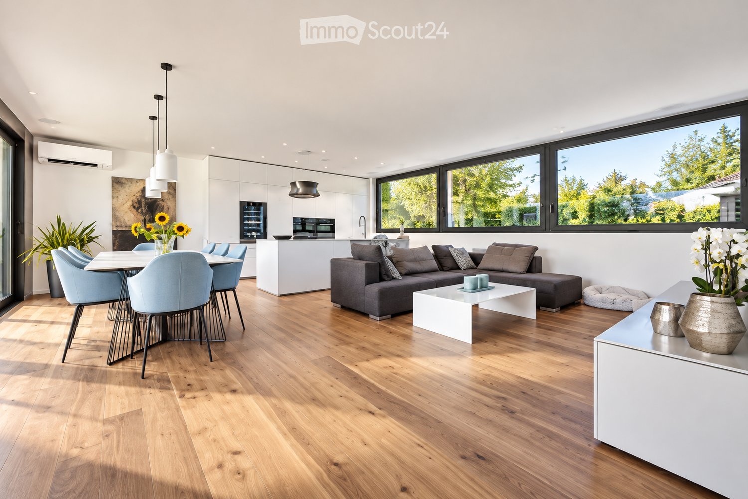 Open-plan living and dining area with light wood flooring, large windows, grey sectional sofa, white coffee table, dining table with chairs, pendant lights, and modern kitchen area.