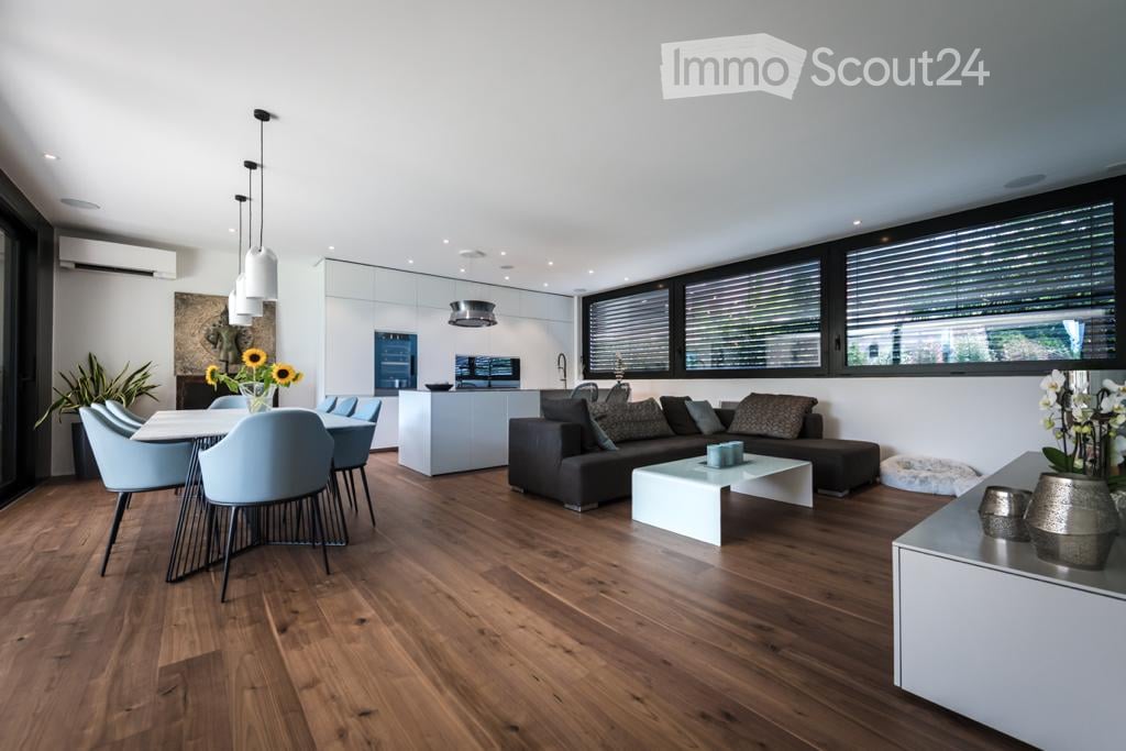 Modern open-plan living and dining area with wooden flooring, large windows, a grey sectional sofa, a white coffee table, a dining table with light blue chairs, pendant lights, and a kitchen island.