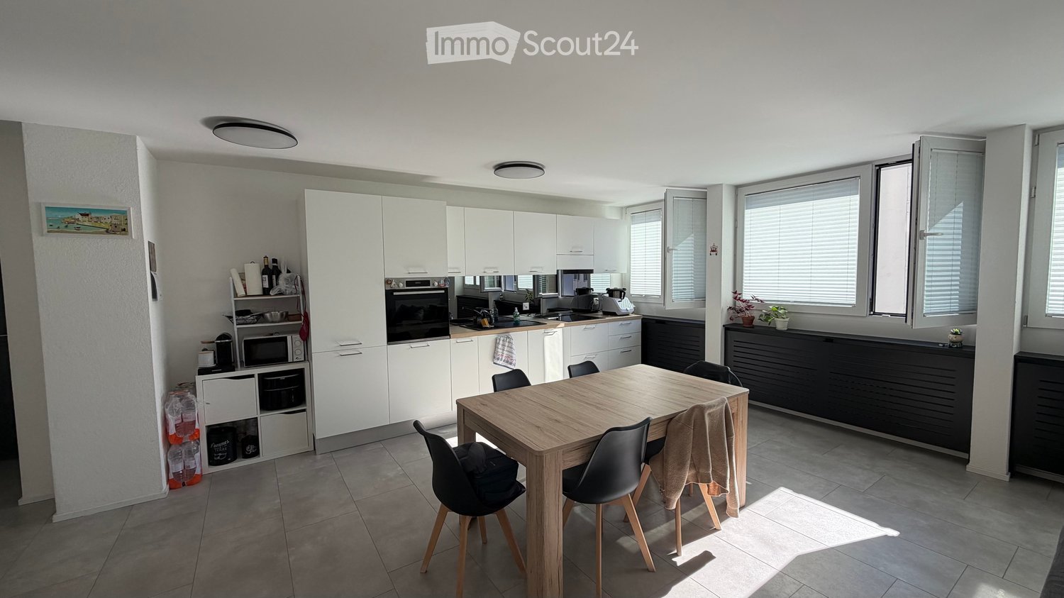 Modern kitchen with white cabinets, a wooden dining table with four chairs, and various appliances including an oven, microwave, and fridge. The space is well-lit with large windows and has a tiled floor.