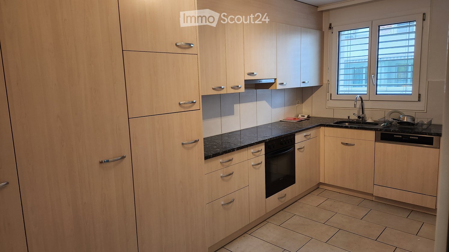 Modern kitchen with light wooden cabinets, black granite countertop, stainless steel sink and faucet, built-in oven and dishwasher, and tiled floor.