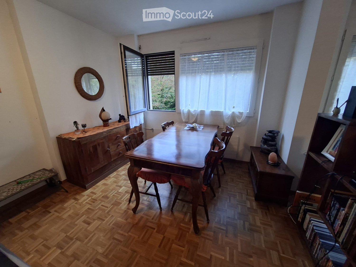 Dining room with wooden floor, large dining table, chairs, cabinet, mirror, and shelves.