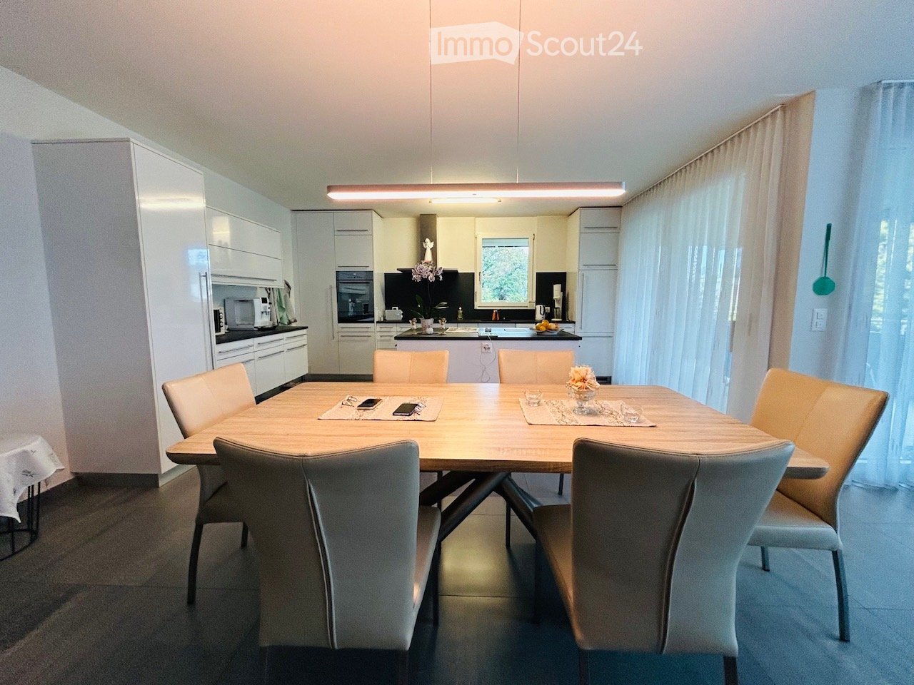 Modern dining room with a wooden dining table surrounded by six chairs, white cabinets, and a small kitchen area with appliances.