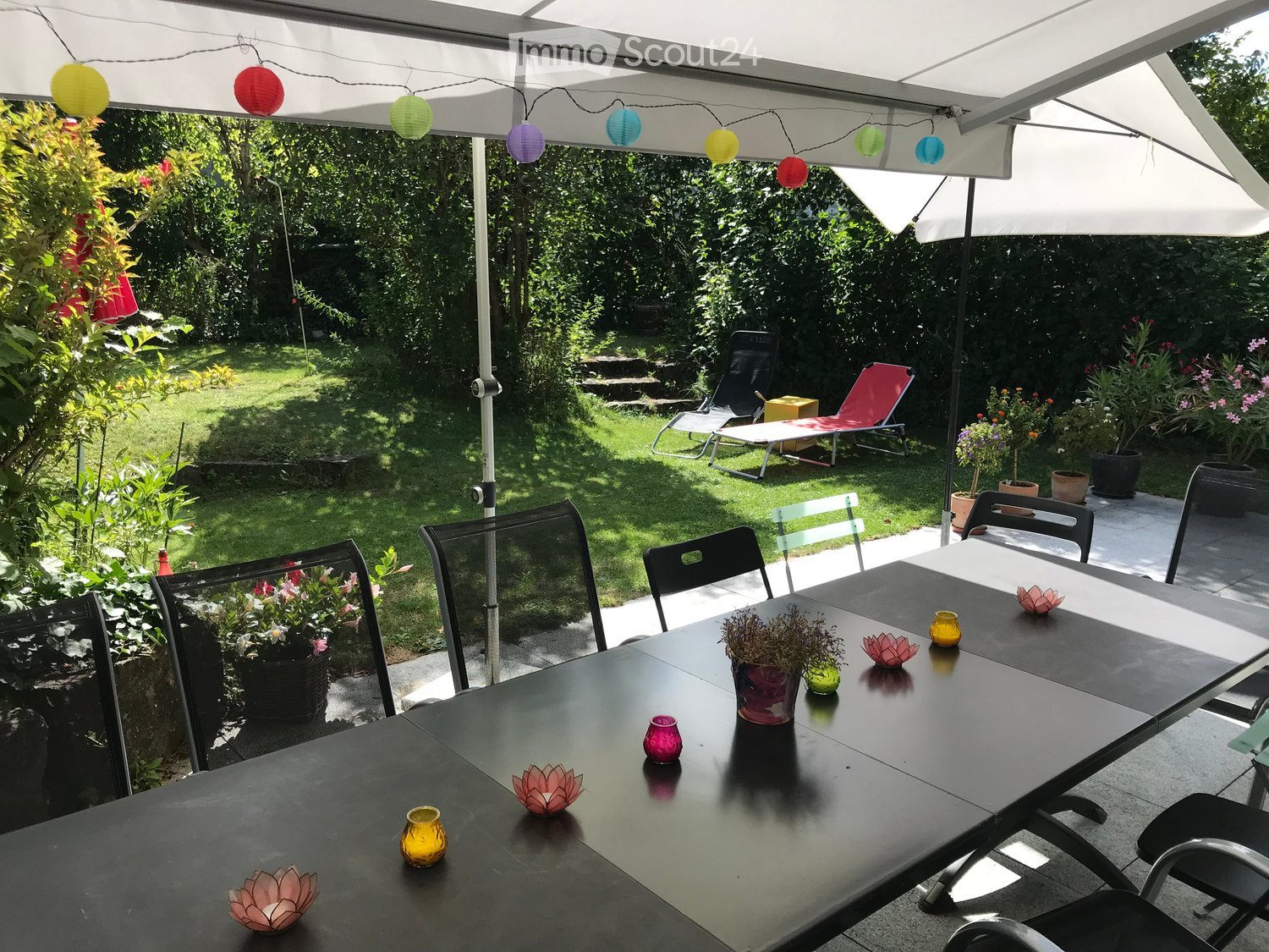 A covered outdoor patio with a black dining table, chairs, and a large umbrella. The area is decorated with small flower pots and candles, with a grassy garden and lounge chairs visible in the background.