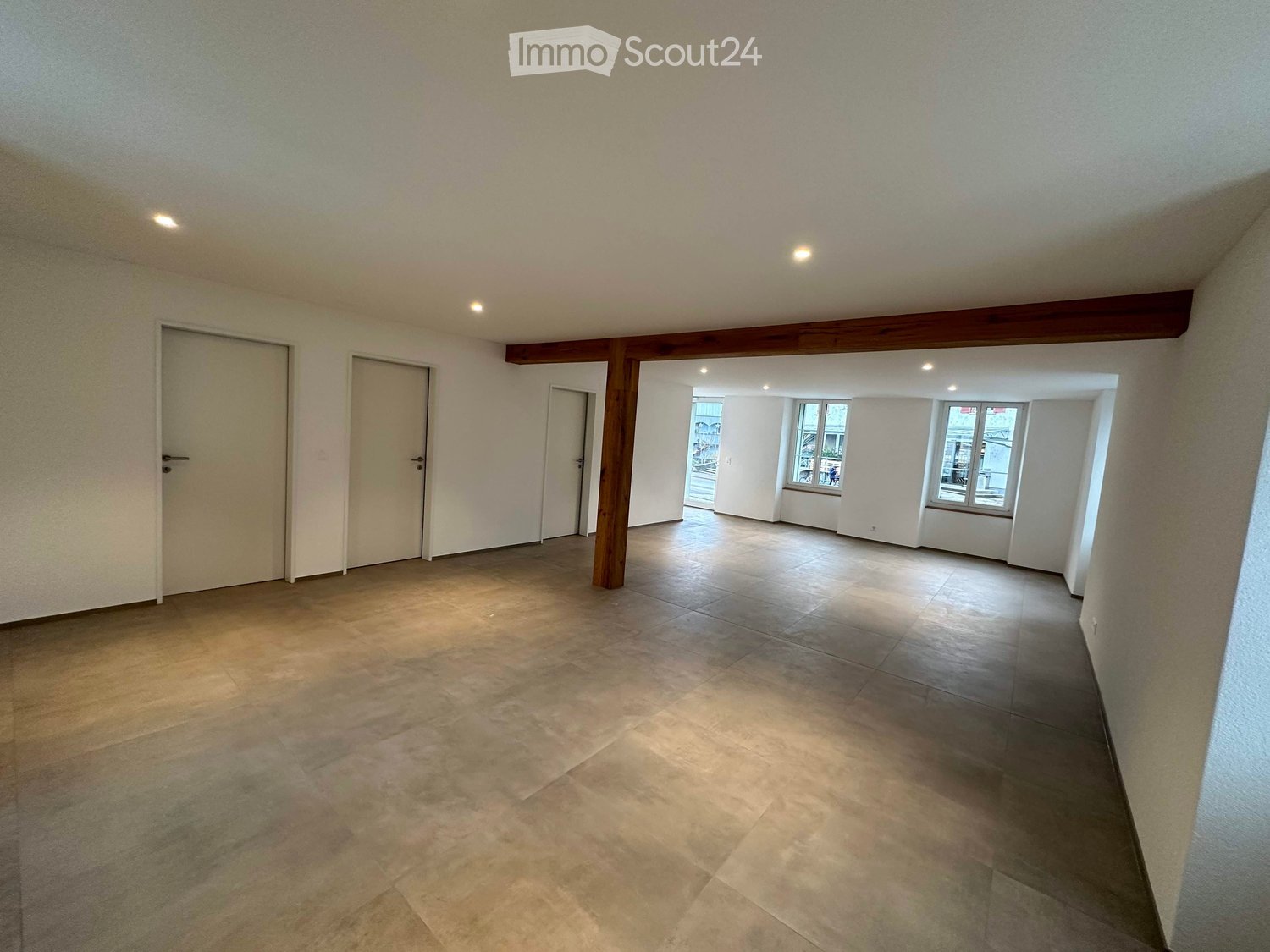Empty room with tiled floor, white walls, and several doors leading to other spaces. Natural light comes through windows in the adjacent area.