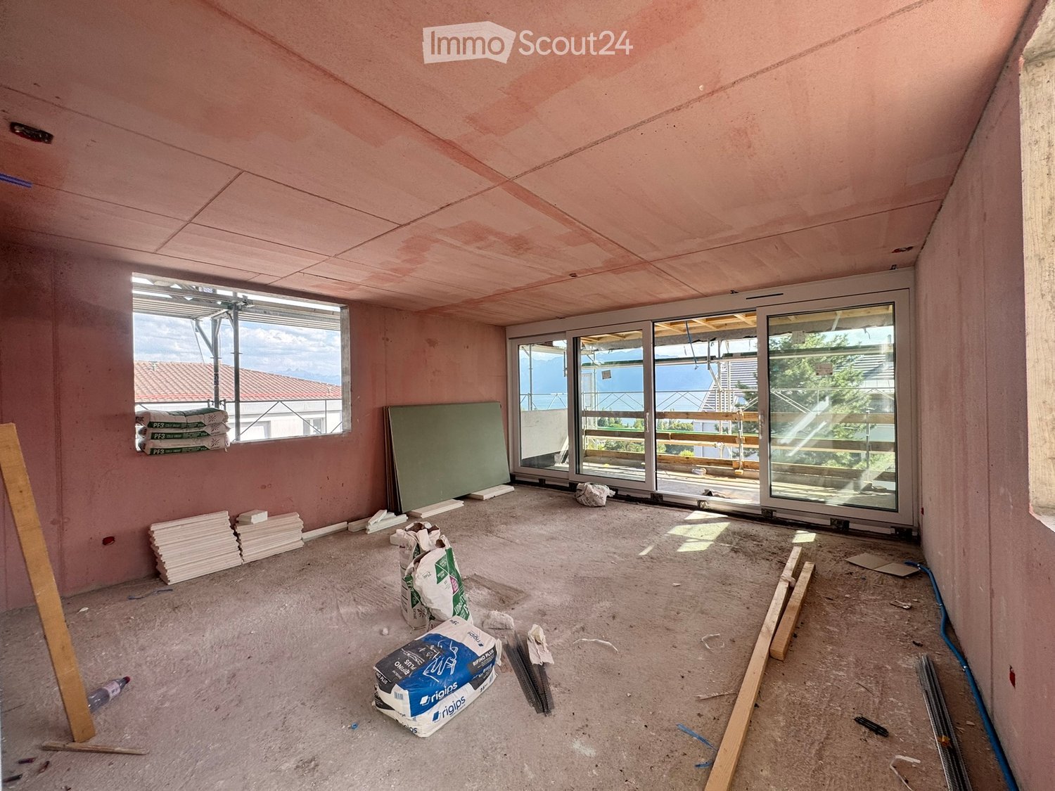 Empty room with unfinished walls and floor, large windows, and construction materials scattered around.