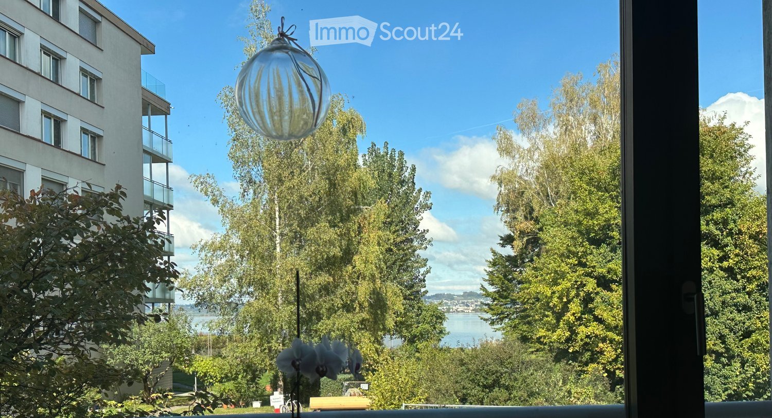 Balcony with a view of a modern apartment building, trees, and a body of water. A hanging glass balloon and a flower arrangement are visible.