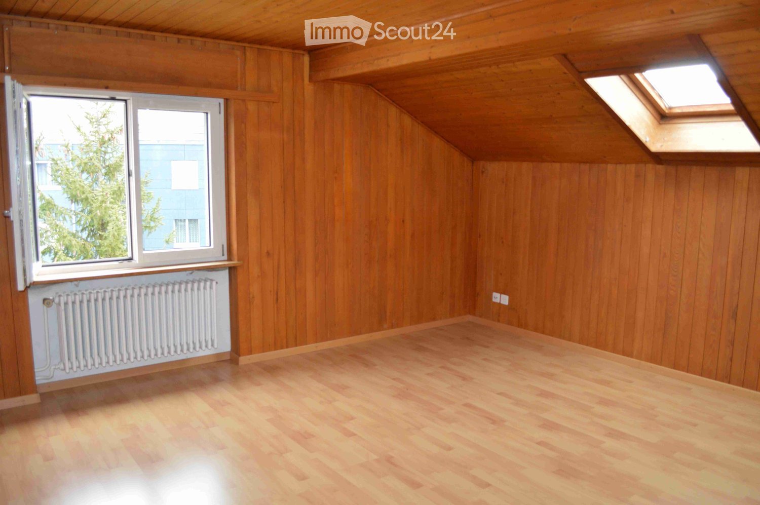 Empty attic room with wooden walls and floor, large window, skylight, and radiator.