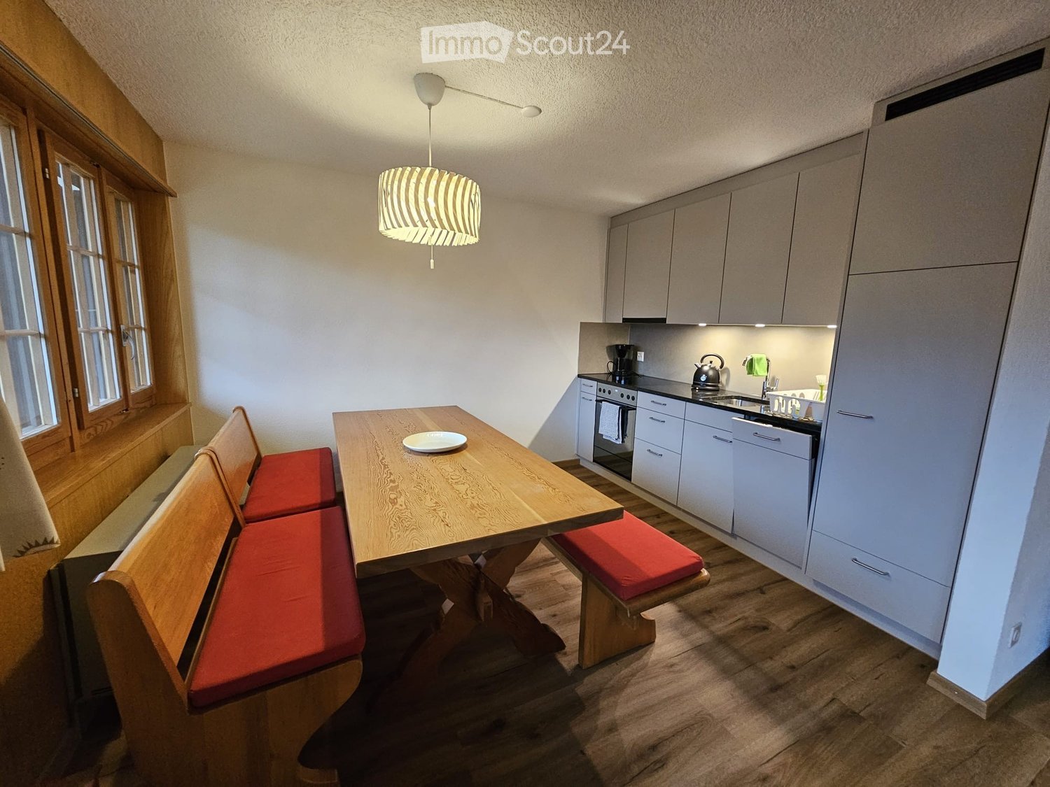 A cozy kitchen and dining area with a wooden table, red cushioned benches, modern white cabinets, and a small kitchen setup including an oven, fridge, and sink.