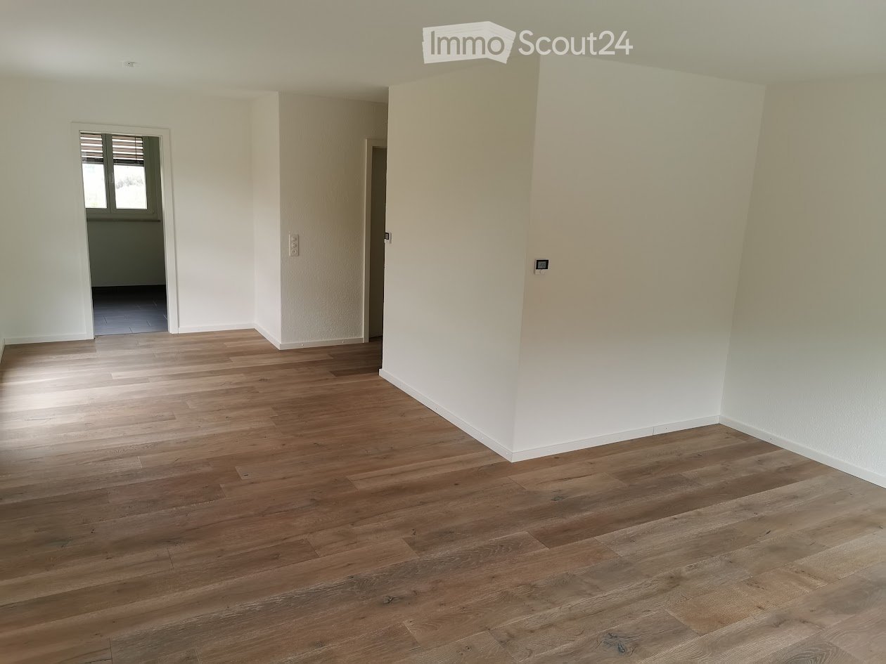 Empty room with light wooden flooring and white walls, featuring a doorway leading to another room with dark flooring.