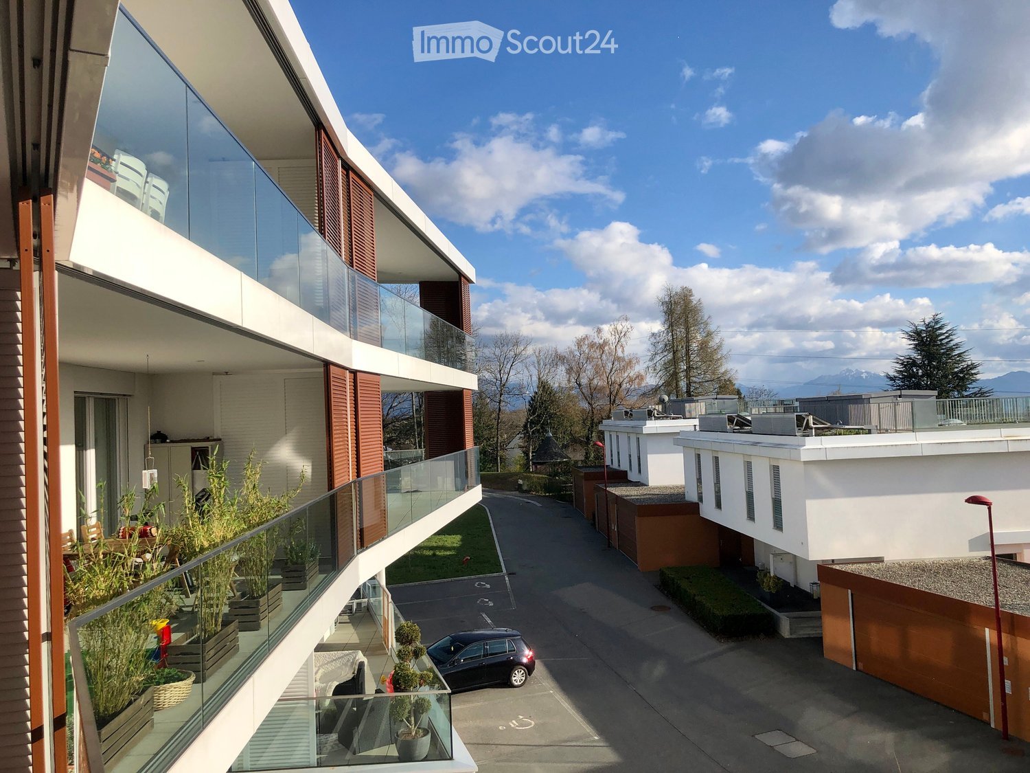 Modern multi-story residential building with balconies, glass railings, and a view of a landscaped courtyard with trees and a parked car.